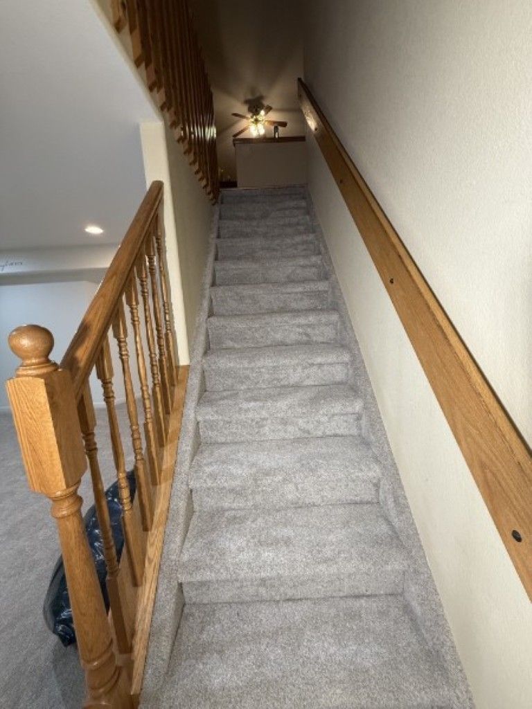 Carpeted staircase with wooden banister leading up to a narrow hallway, viewed from below.