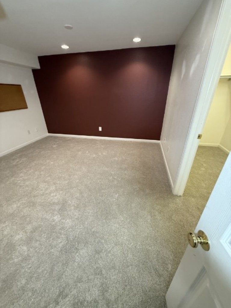 Empty carpeted room with burgundy accent wall, white walls, and open door with hallway beyond
