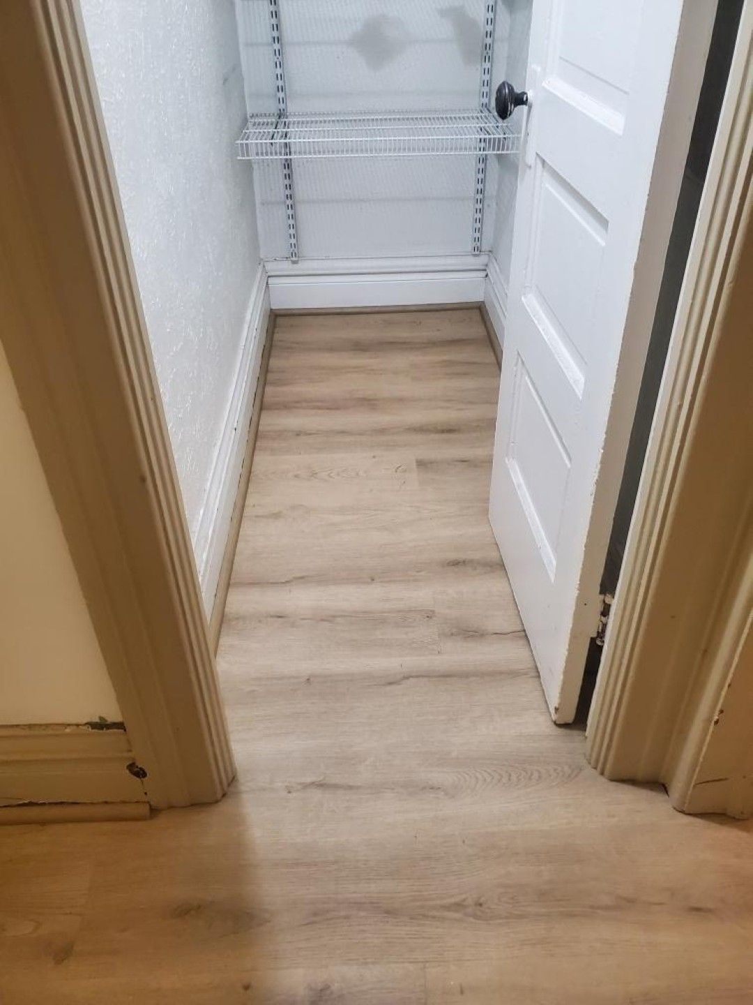 Narrow hallway with light wood floor, white closet shelves, and an open white door.