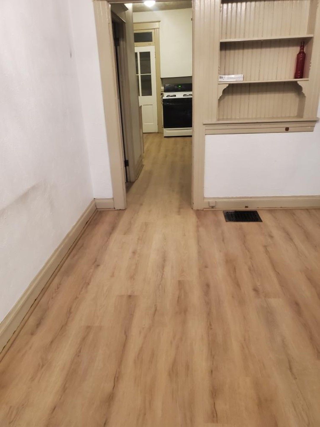 Narrow hallway with light wood flooring, white walls, built-in shelves, and a kitchen visible at the end.