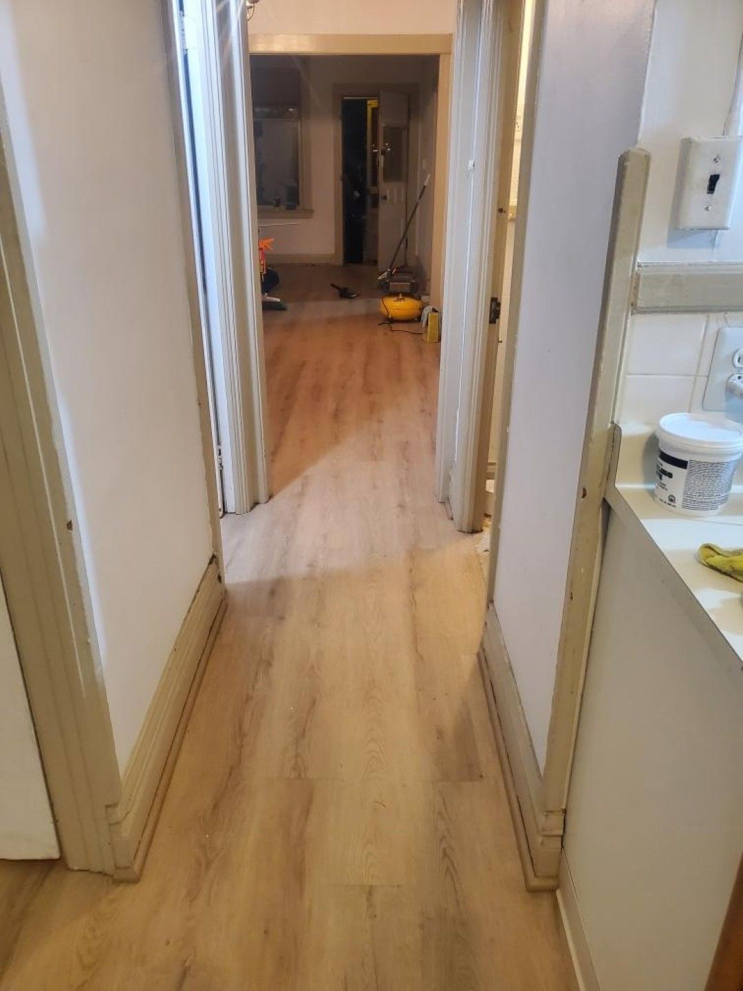 Narrow indoor hallway with wood flooring, leading to a room with scattered items on the floor.