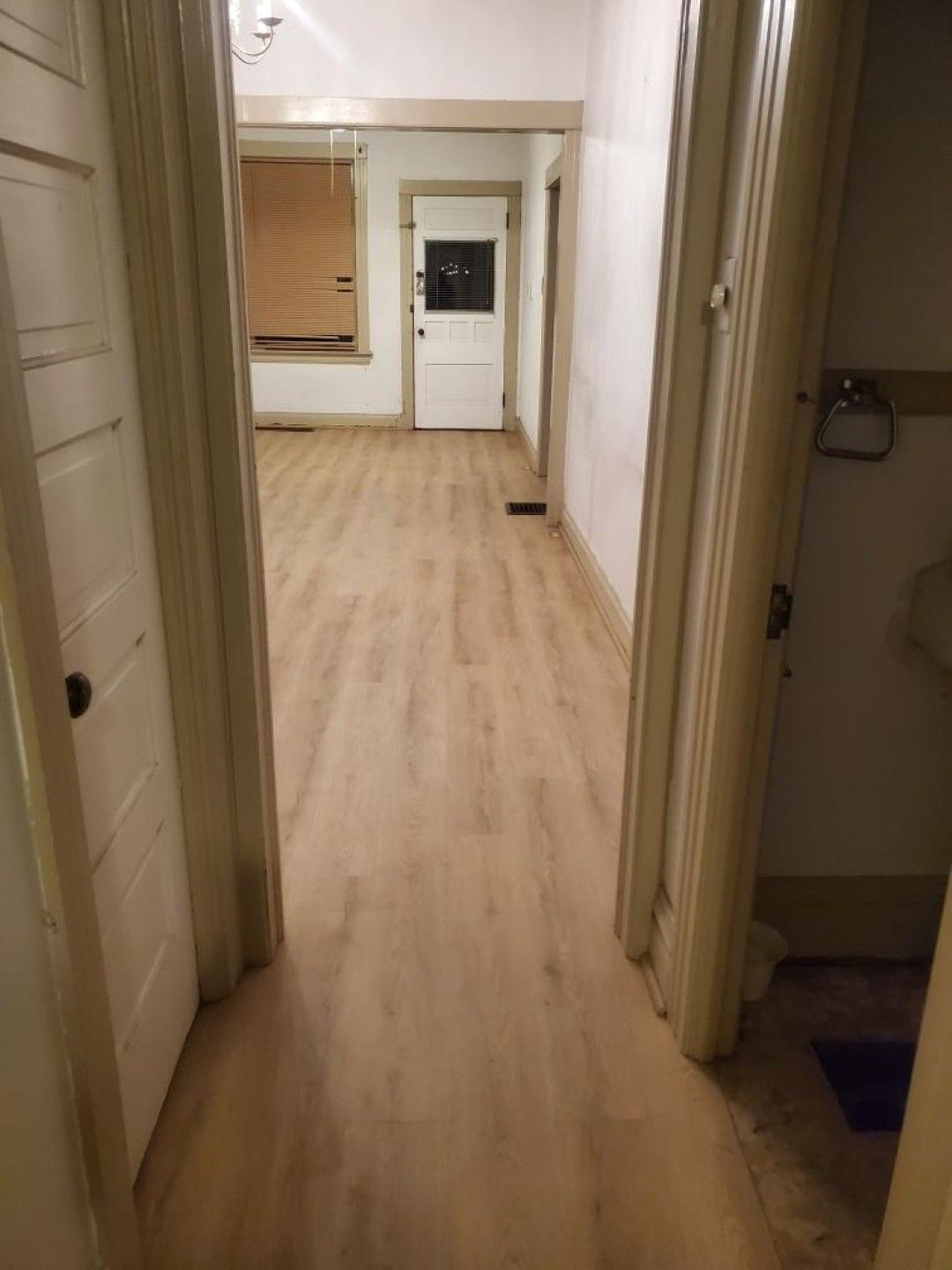 Narrow hallway with light wood flooring, white walls, and doors leading to a small room or entryway.