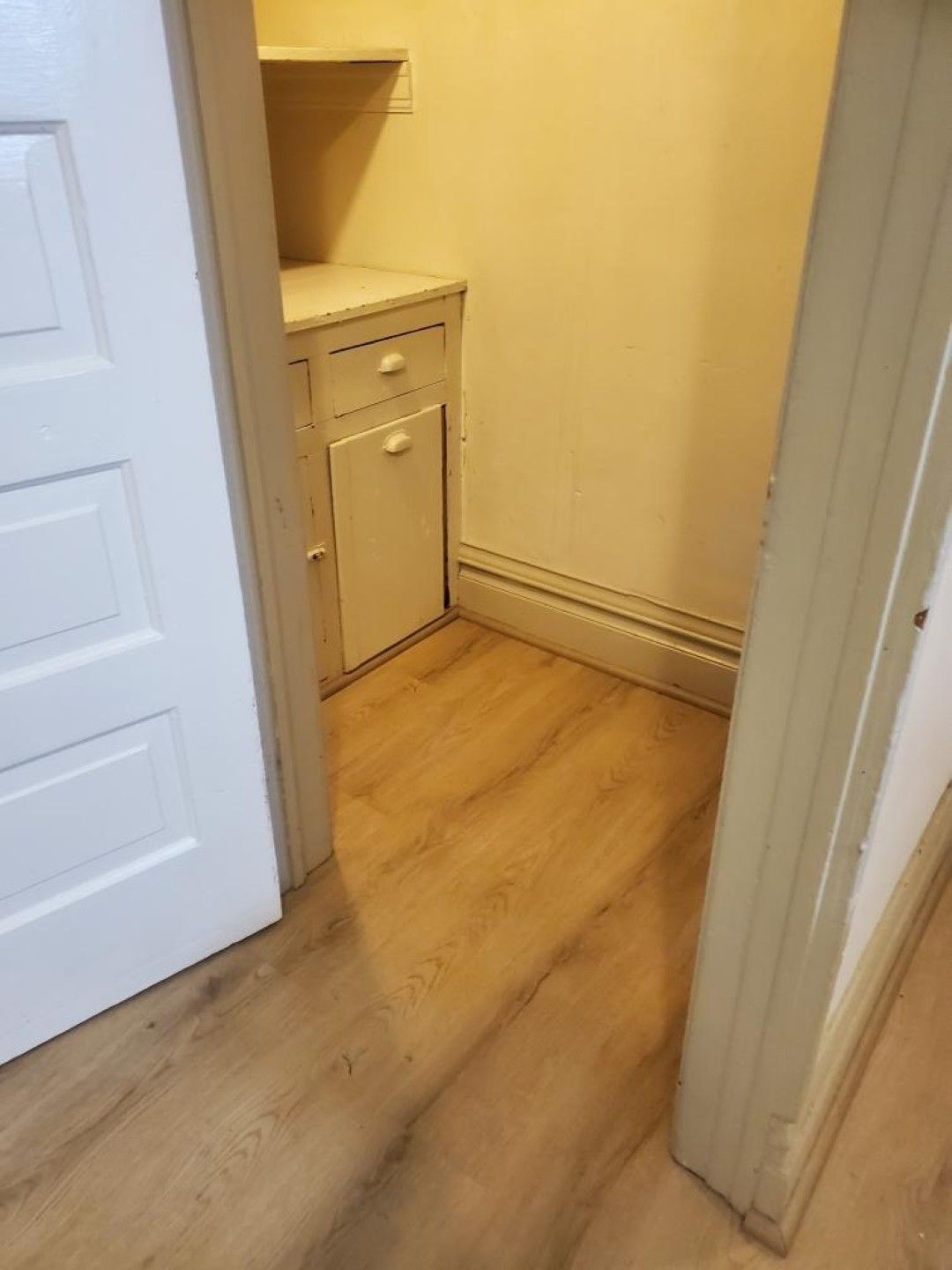 Small hallway with light wood flooring, beige walls, and a compact white cabinet near a doorway.