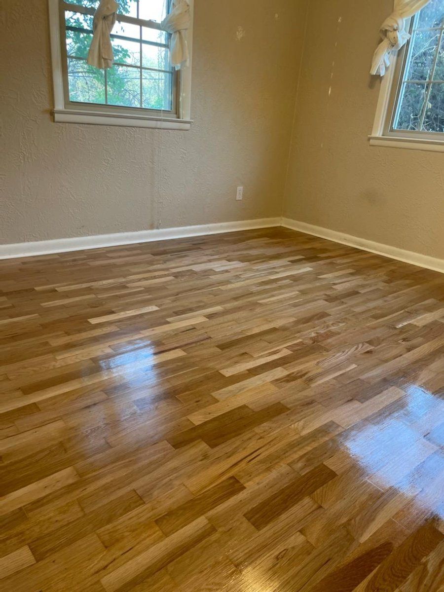 Empty room with glossy hardwood floors, beige walls, and two windows letting in daylight