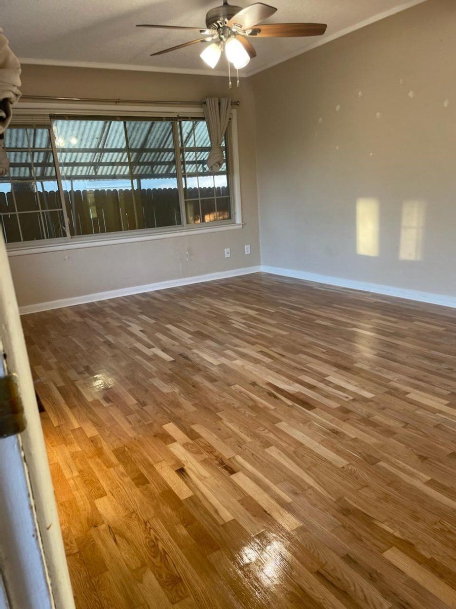 Empty living room with hardwood floors, beige walls, ceiling fan, and large front window.