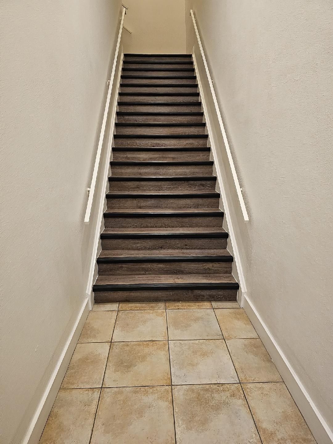 Narrow indoor staircase with dark steps and white walls above a tiled floor corridor