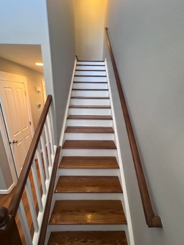 Narrow indoor staircase with wooden treads, white risers, and brown handrails beside a hallway.