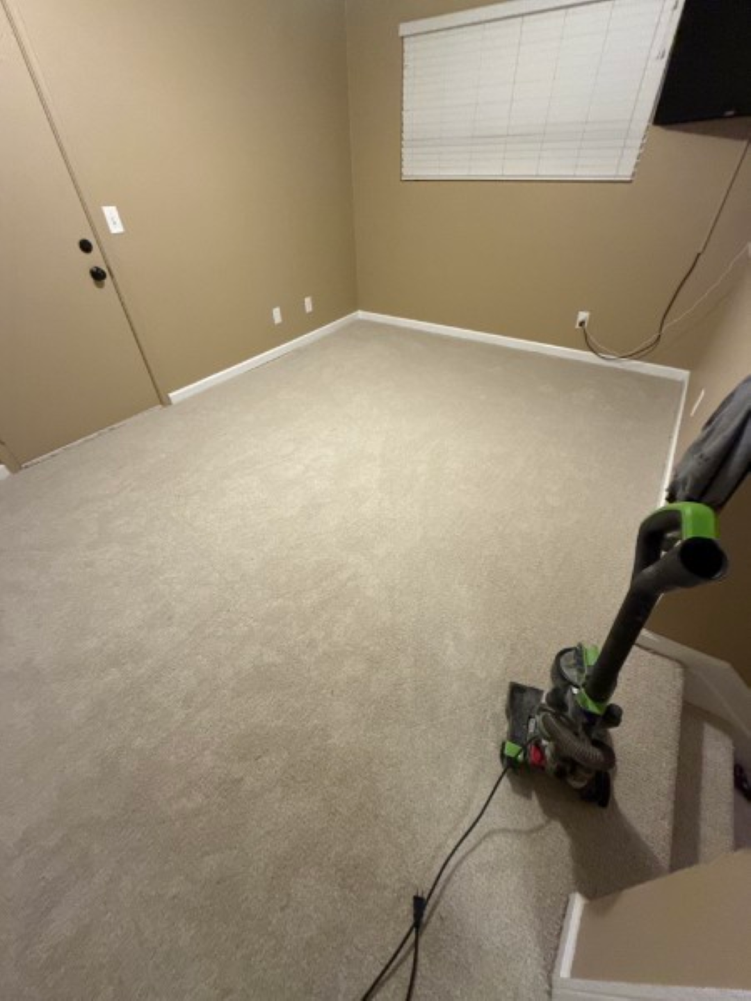 Empty beige bedroom with carpet; a vacuum cleaner sits near the stairs and window.