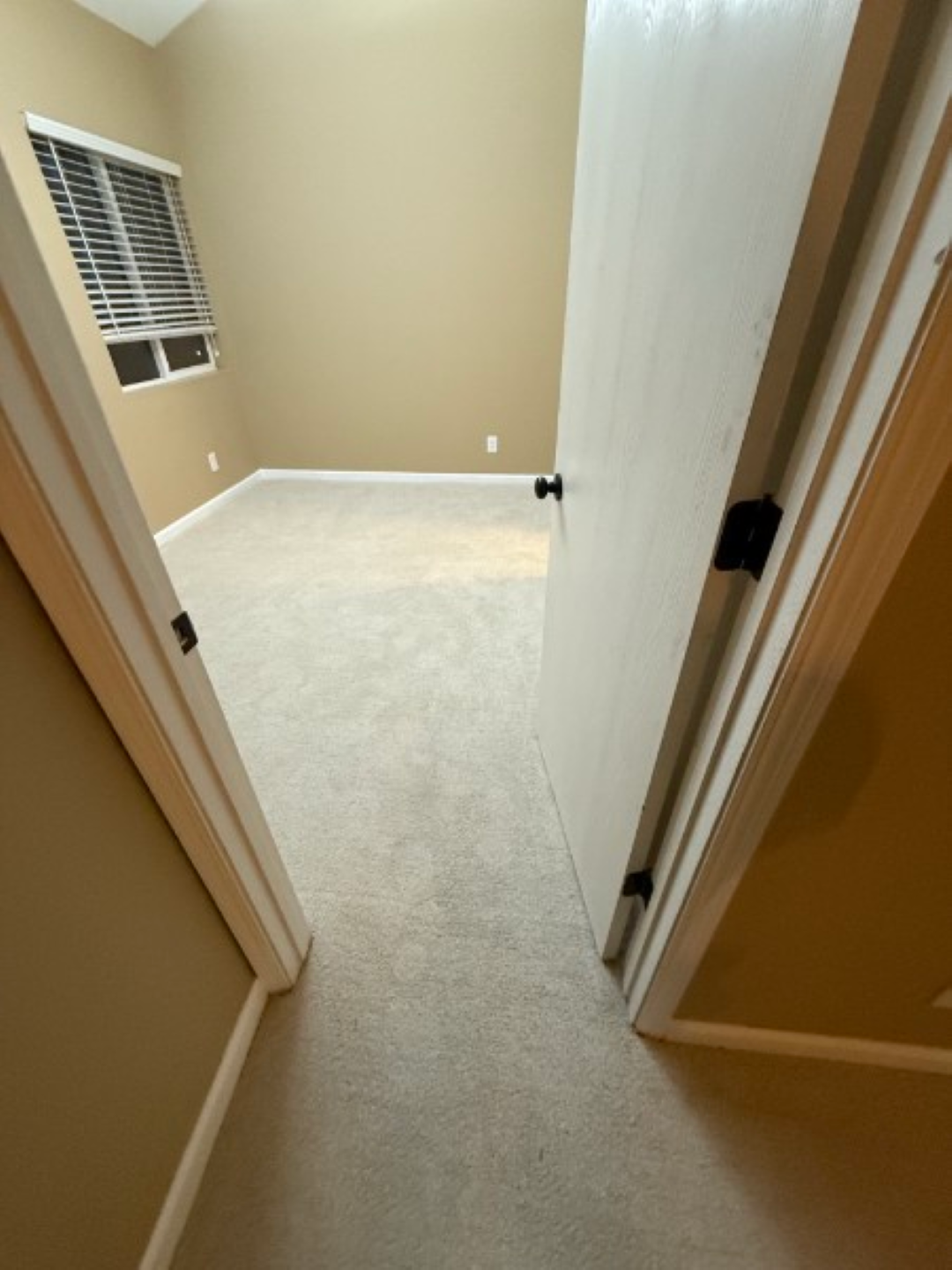 Empty beige bedroom seen through open doorway, with carpeted floor and window blinds on the left.