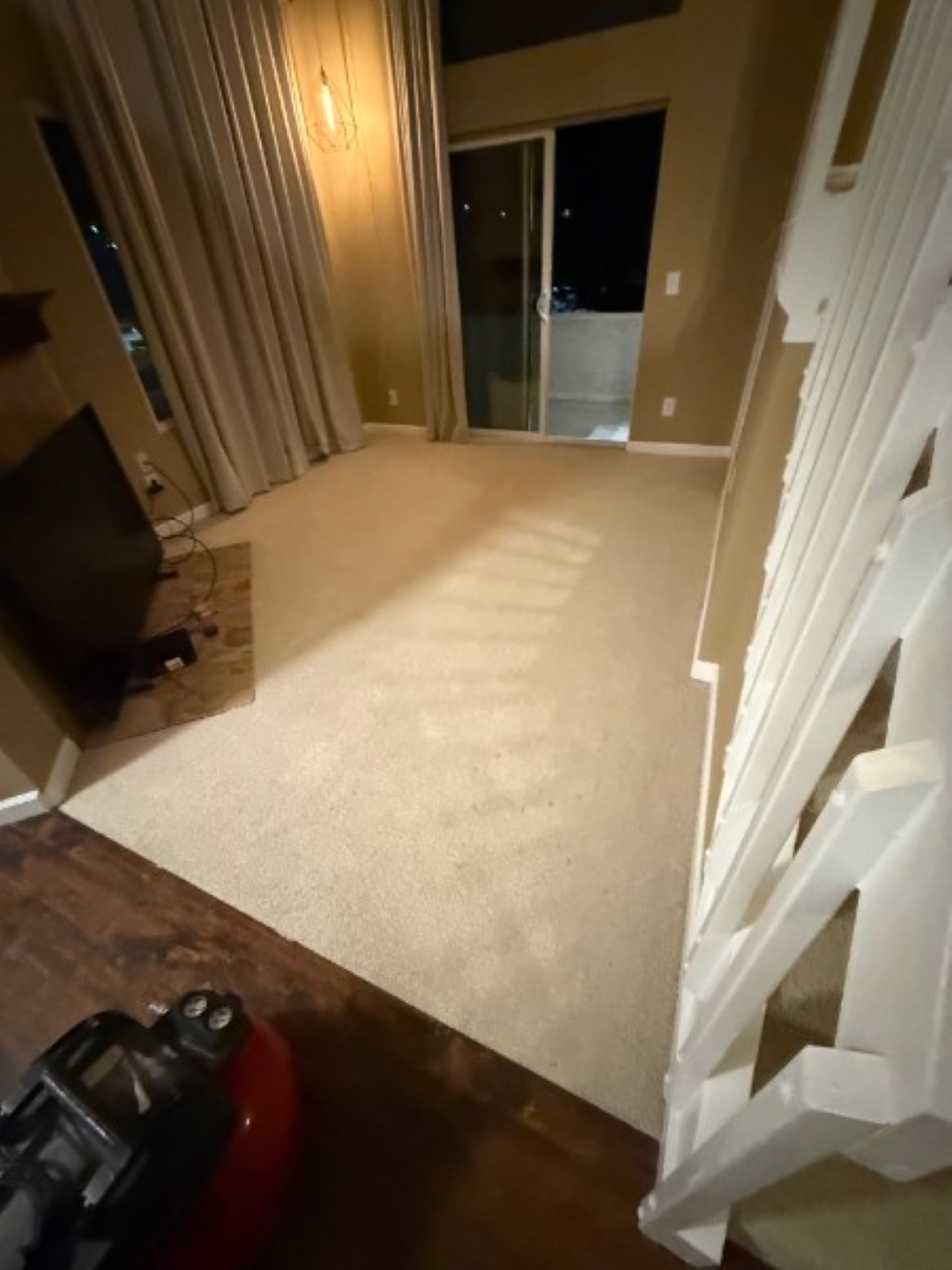 Empty beige carpeted room with sliding glass doors and draped curtains, lit by warm indoor lighting