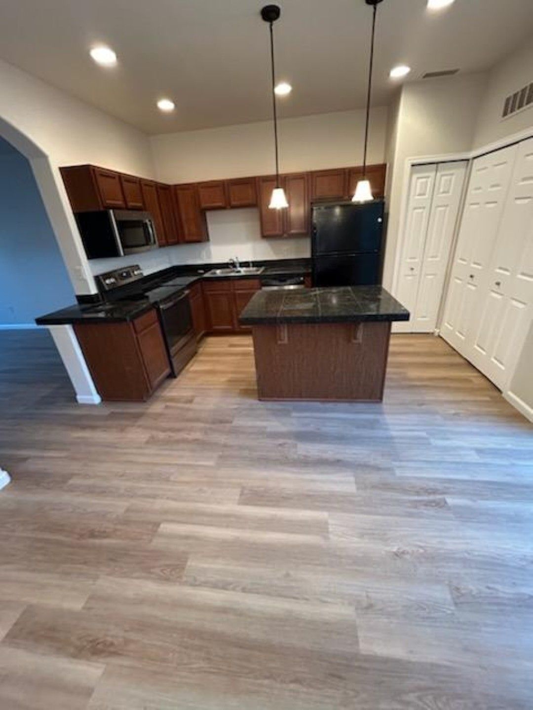 Empty modern kitchen with dark wood cabinets, black counters, island, pendant lights, and light wood flooring