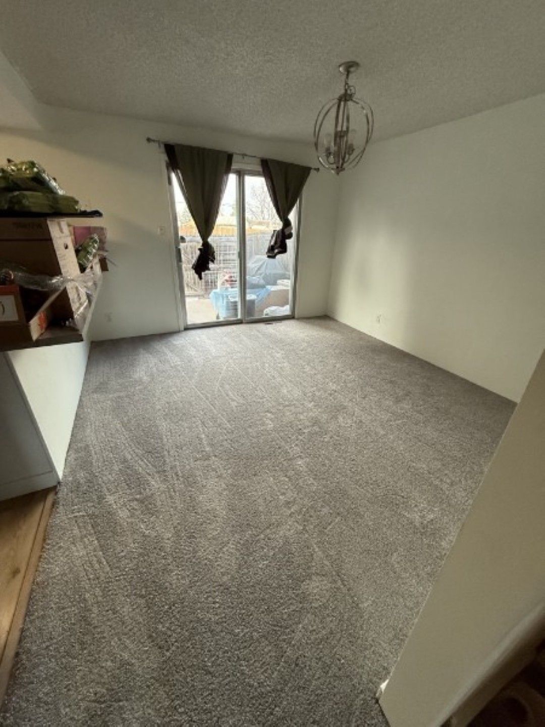 Empty carpeted room with white walls, a hanging light fixture, and a sliding glass door with dark curtains.