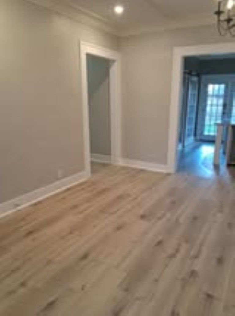 Empty modern living room with light wood floors, white walls, recessed lights, and a doorway to another room