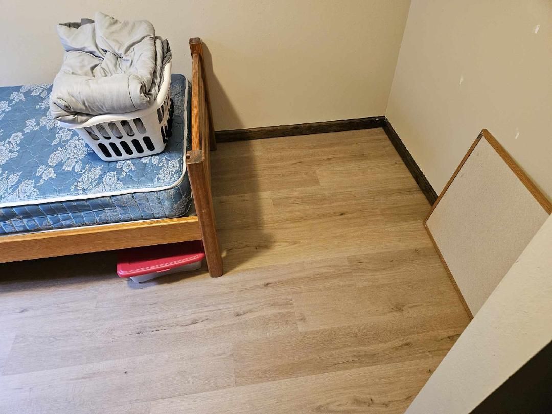 Small bedroom corner with a bed, laundry basket, and empty floor space against beige walls.