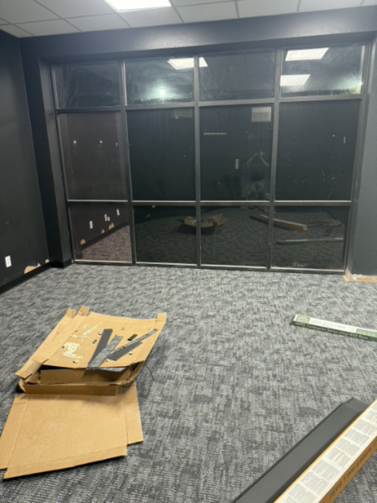 Empty office with glass wall, carpet tiles, and cardboard boxes and packaging debris on the floor.