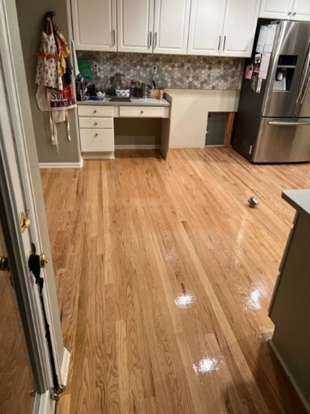 Bright kitchen with white cabinets, wood floor, stainless fridge, and an empty desk area.