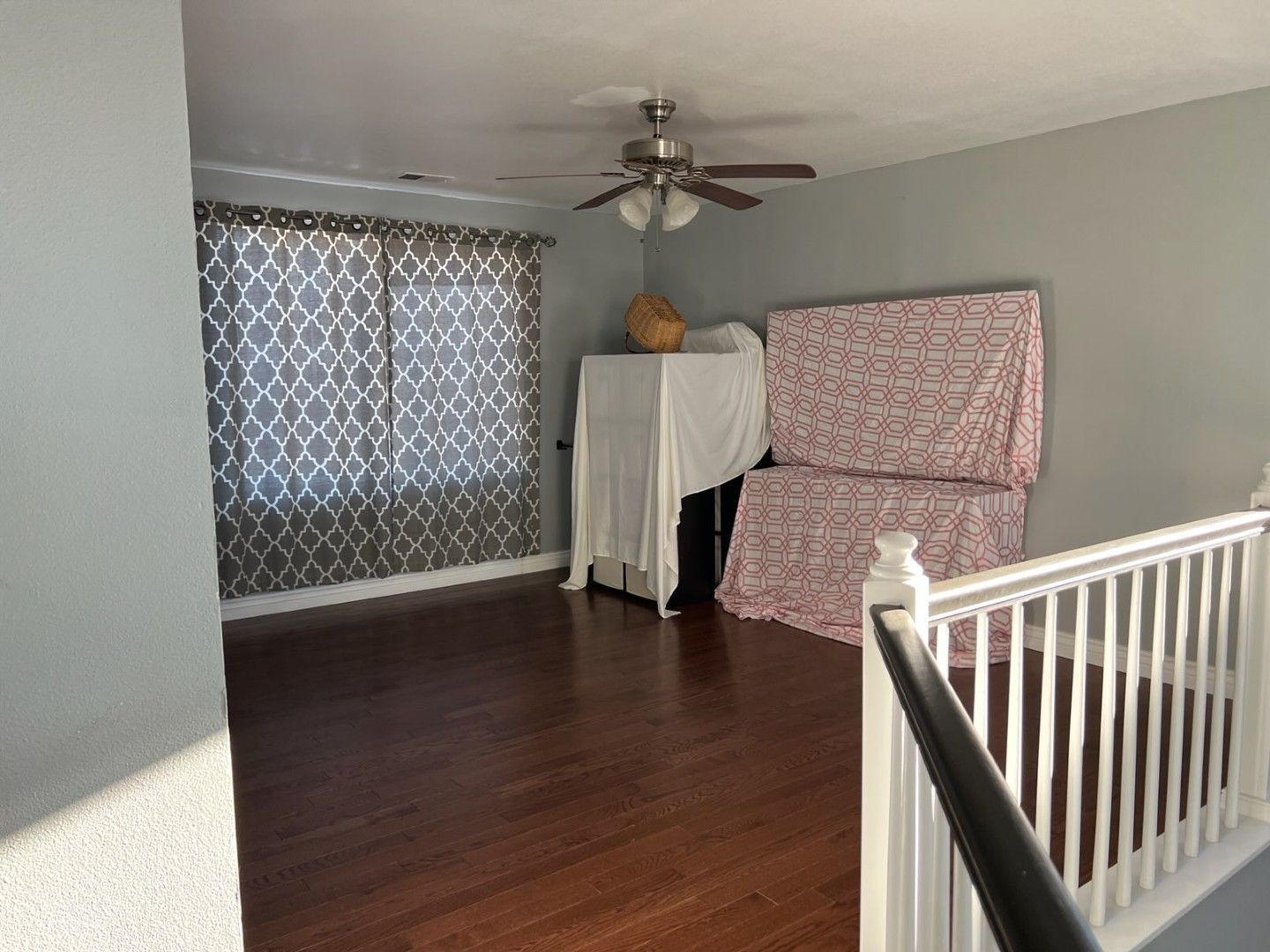 Empty room with hardwood floor, ceiling fan, and covered furniture near a window.