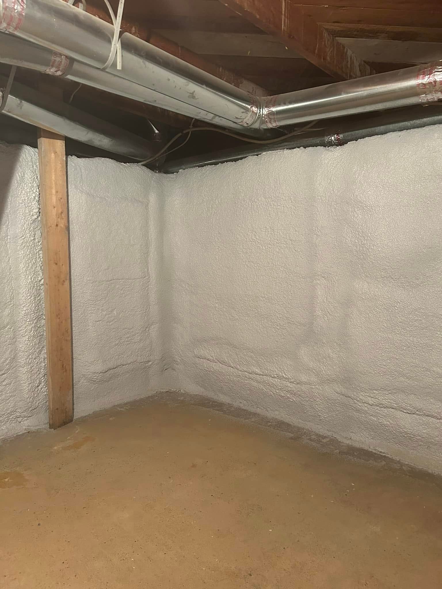 An unfinished basement corner with spray foam insulation on concrete walls and a wooden support beam.