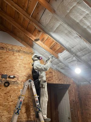 A worker in full protective gear stands on a ladder, spray-coating the underside of a wooden roof deck with insulation.