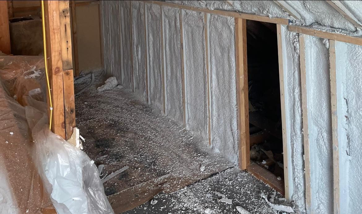 An unfinished attic space with spray foam insulation on the walls, a wooden frame, and construction debris on the floor.