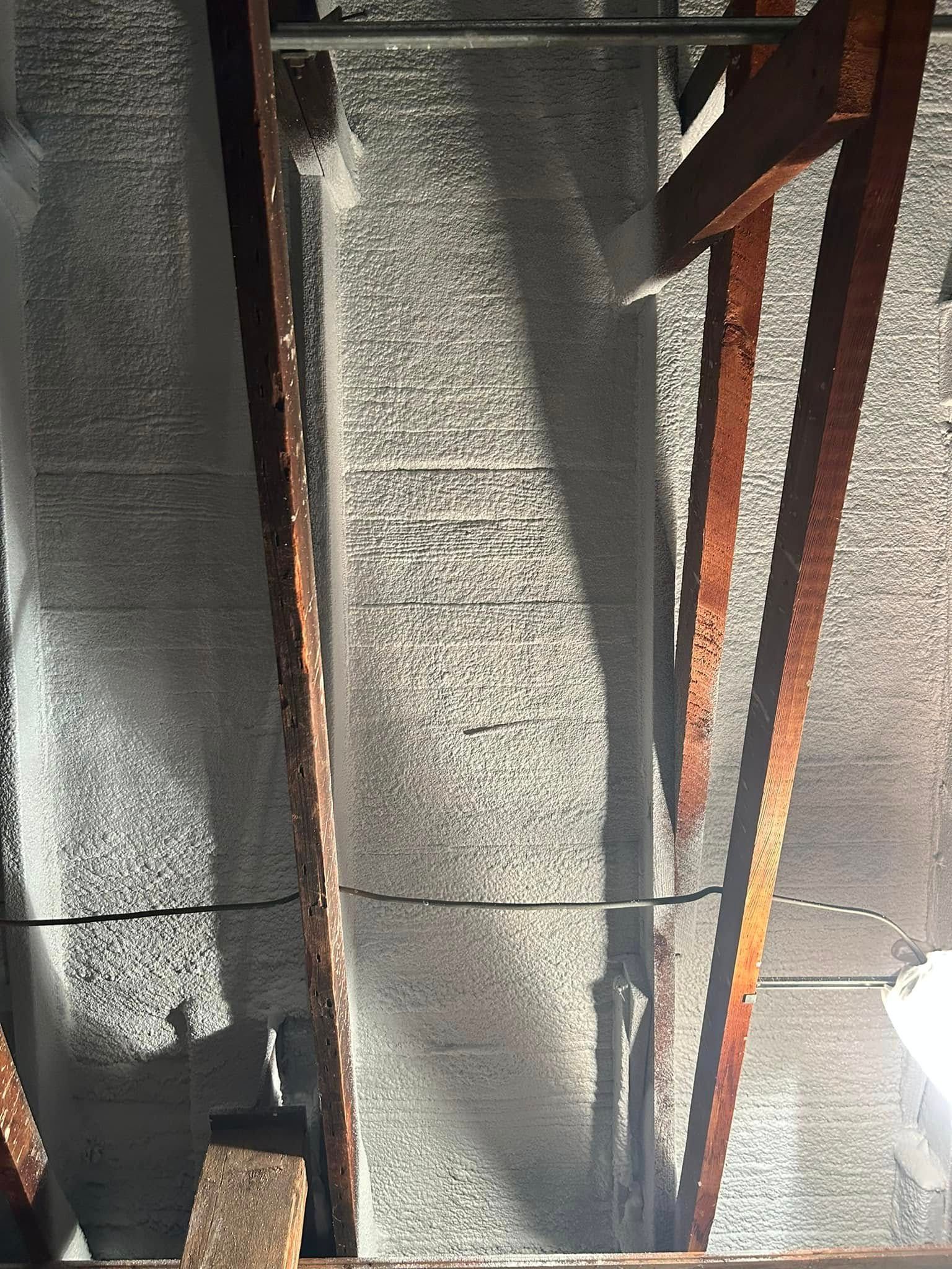 Spray foam insulation applied between wooden roof rafters, with a dark wire running horizontally across the beams.
