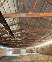 Interior of a large barn with wooden roof trusses, exposed insulation panels, brick walls, and a dirt floor.