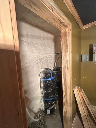 A utility closet containing two dark-colored water treatment tanks set against a white, textured insulated wall.