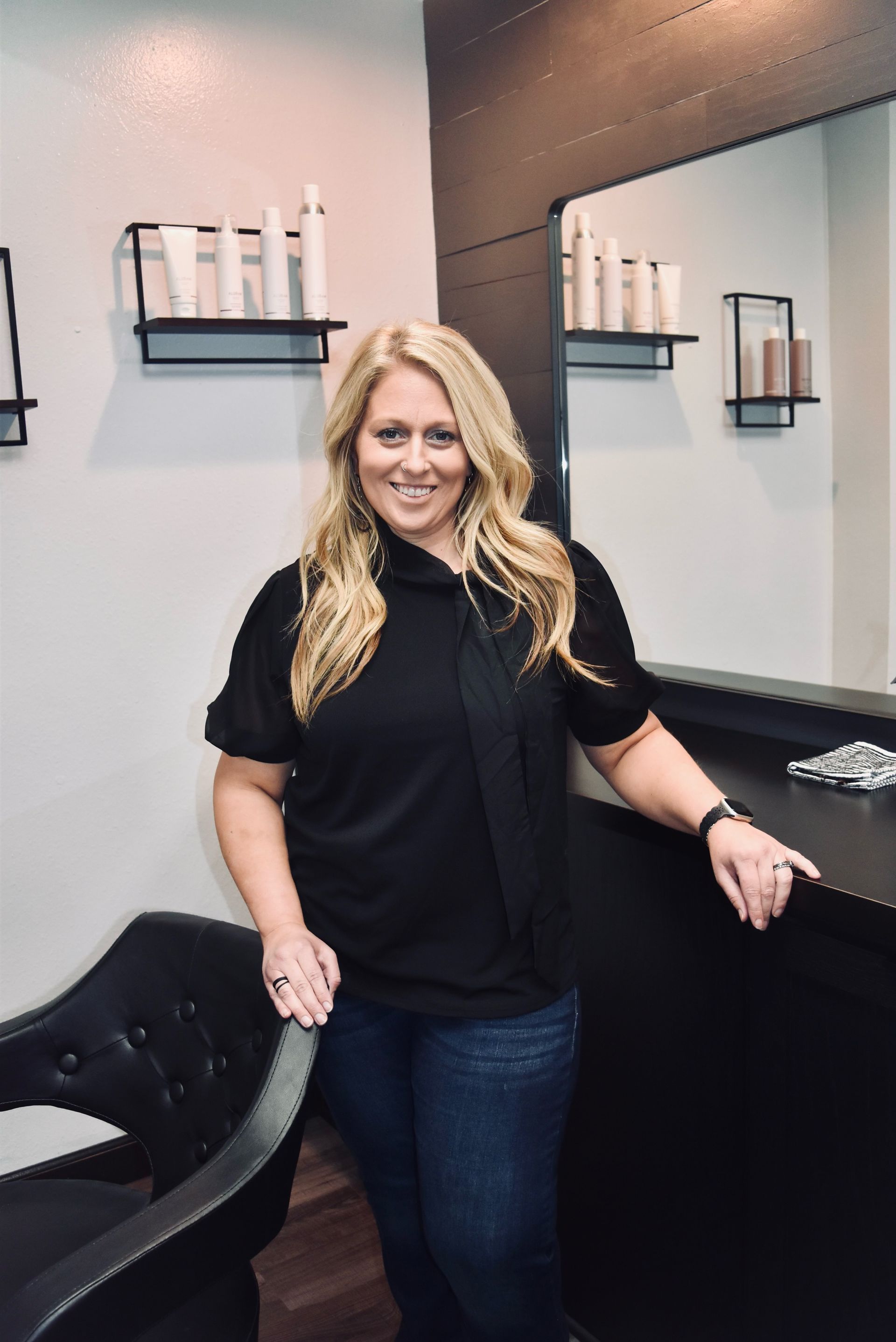 A woman in a black shirt is standing next to a chair in a salon.