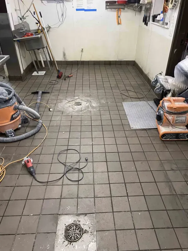 A commercial kitchen under repair with brown tiled floors, two floor drains, a shop vacuum, and tools scattered around.