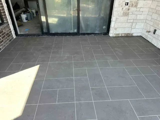 A grey stone patio with a modular paver pattern leads up to a sliding glass door and a white stone exterior wall.