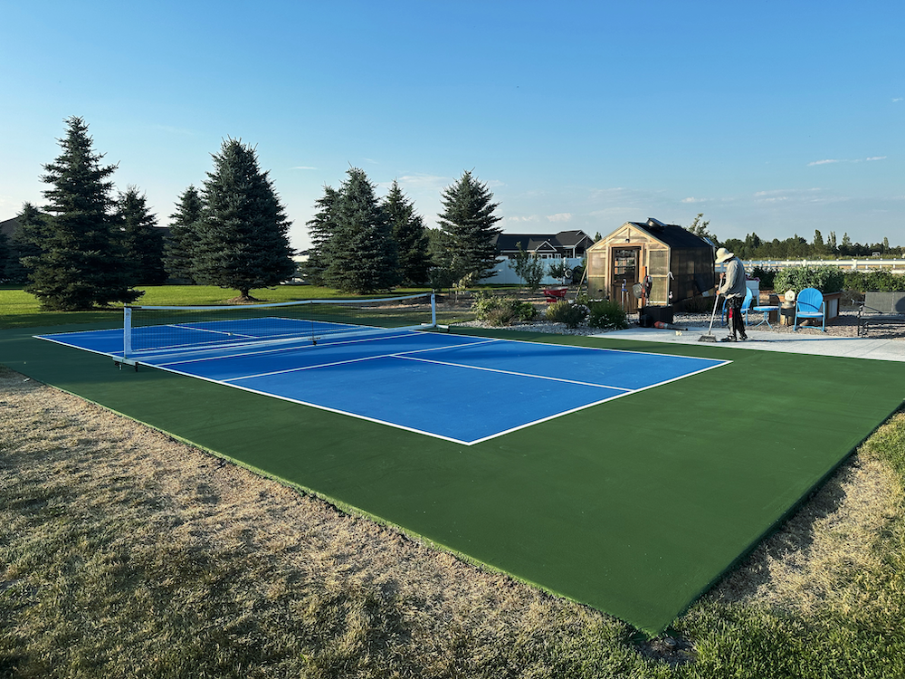 A newly built backyard pickleball court with a blue playing surface and green surrounding area, near a wooden shed.