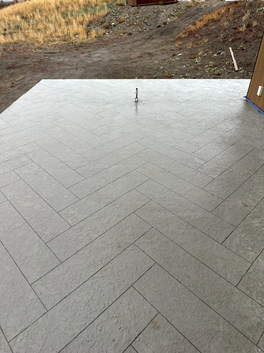 A rectangular concrete patio with a herringbone pattern finish, outdoors against a dirt and grass landscape.