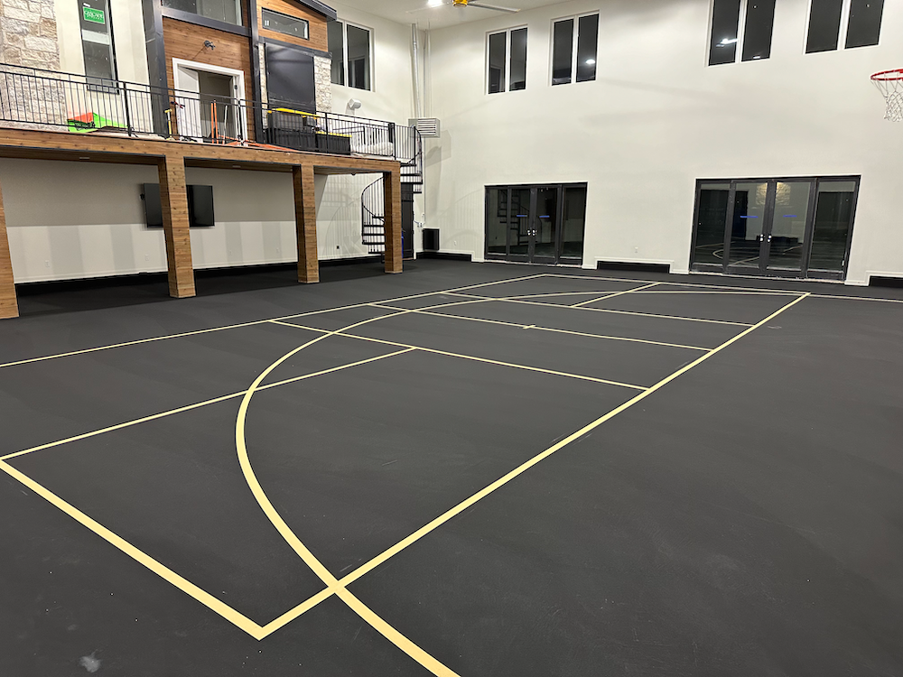 Indoor basketball court with a matte black floor and yellow painted lines, featuring a wooden mezzanine and white walls.
