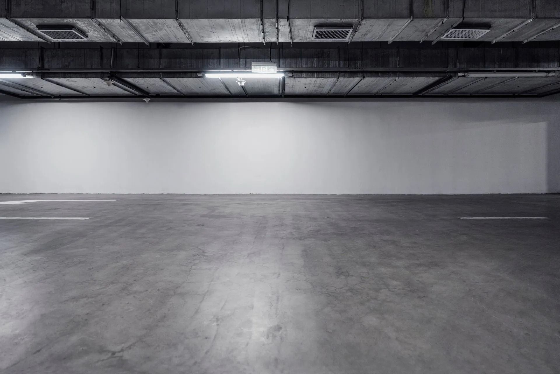 An empty underground parking garage featuring a gray concrete floor, a plain white wall, and industrial ceiling lights.
