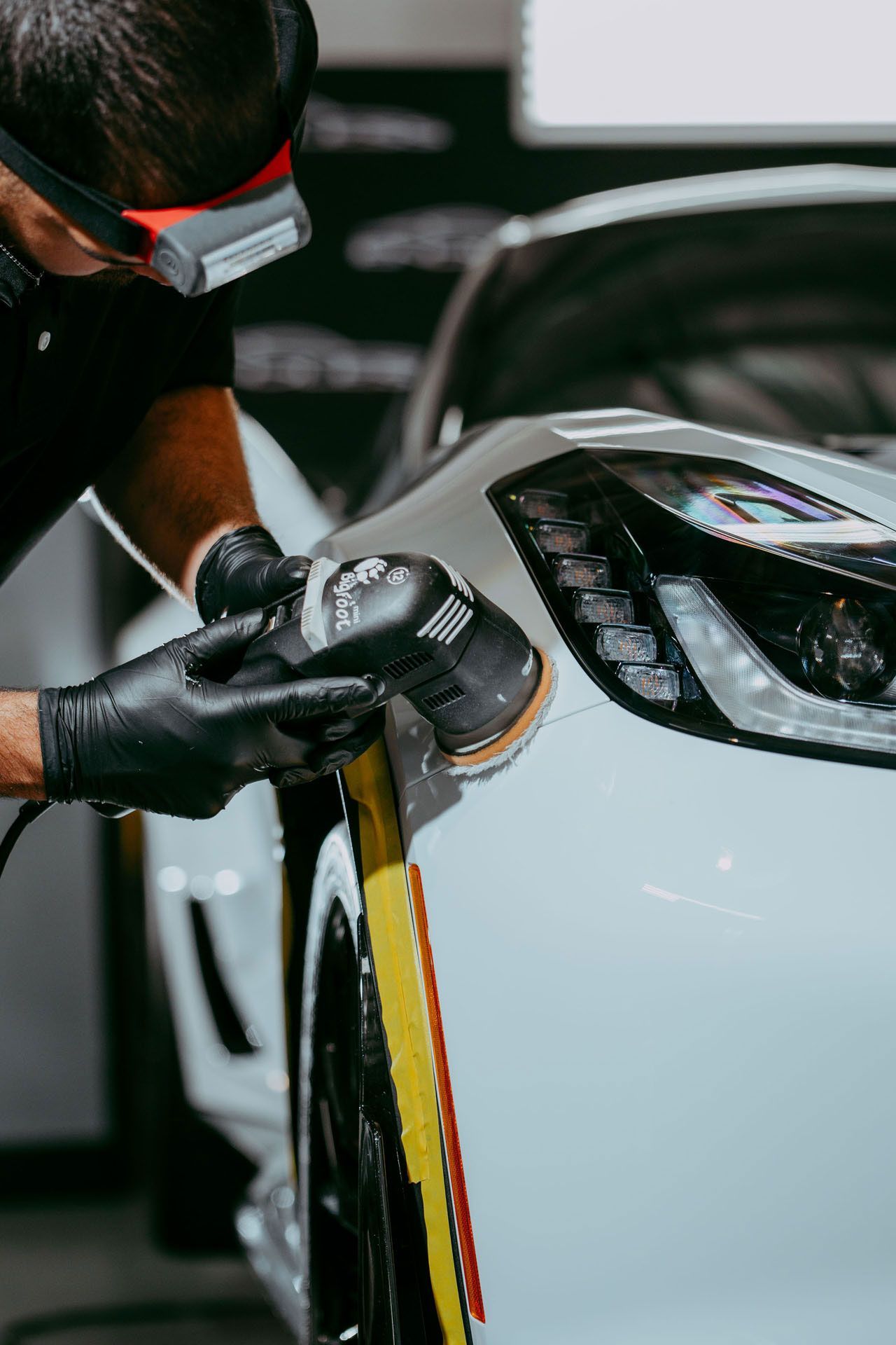 Person polishing white car with power tool, wearing safety glasses and gloves.