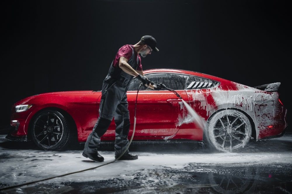 Man washing a red sports car with a pressure washer, surrounded by foam, dark setting.