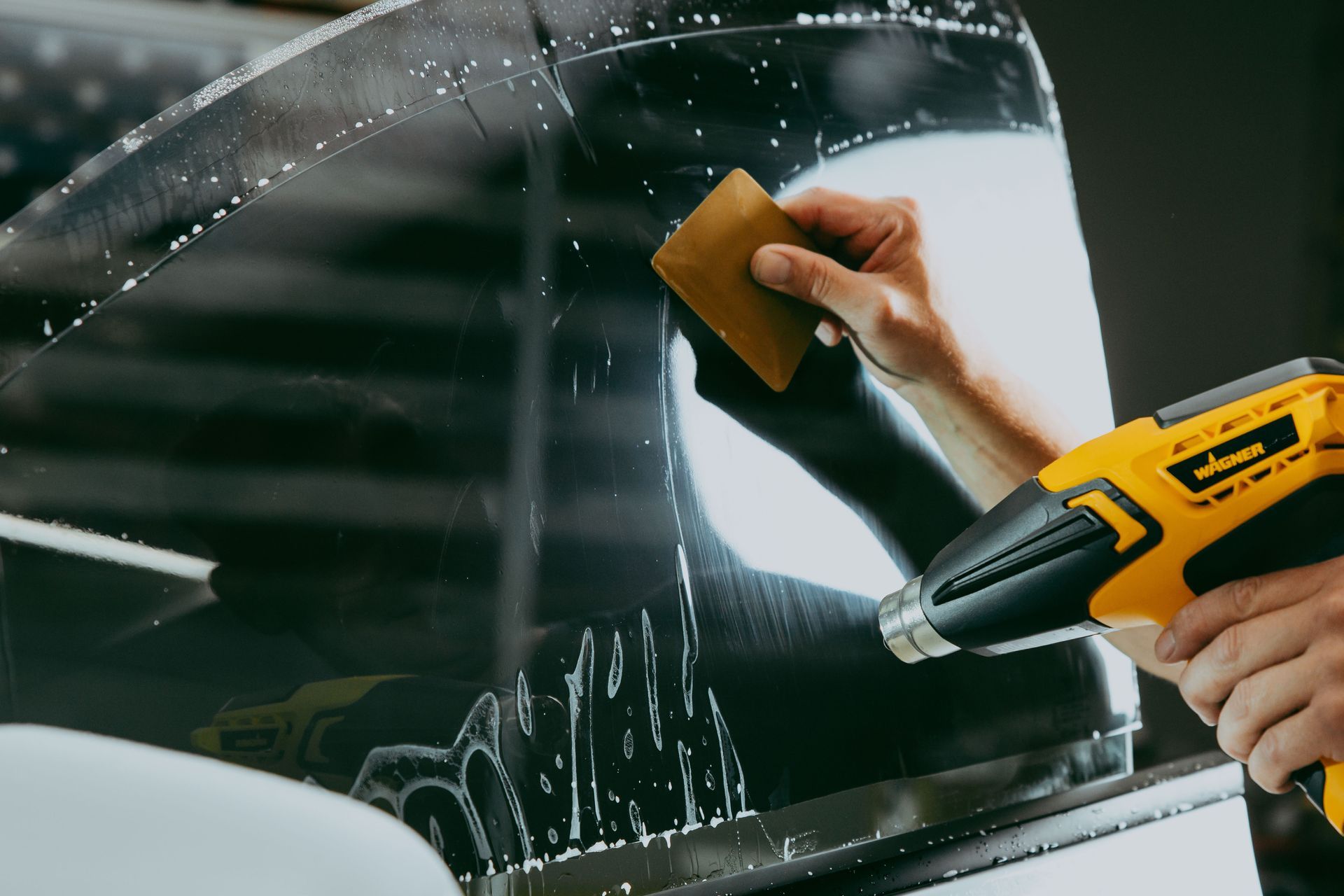 Person applying window tint to a car window, using a squeegee and heat gun.
