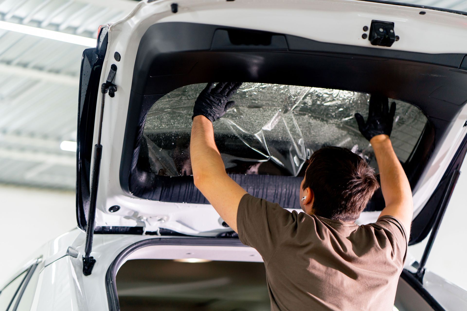 Person installing window tint on a car's rear window. The person wears gloves and is inside a well-lit space.