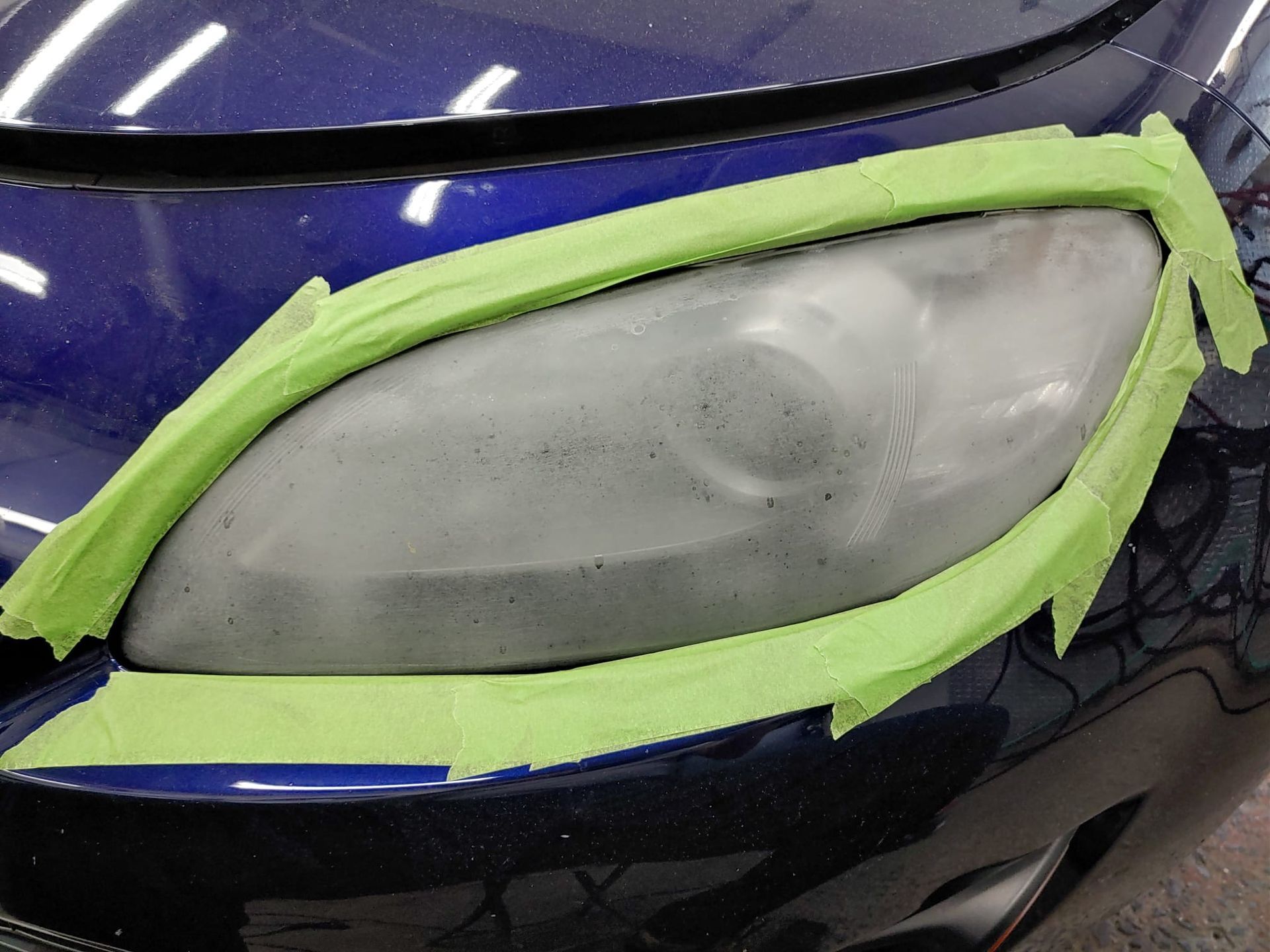 A car headlight covered in green tape, being prepared for restoration against a blue car body.