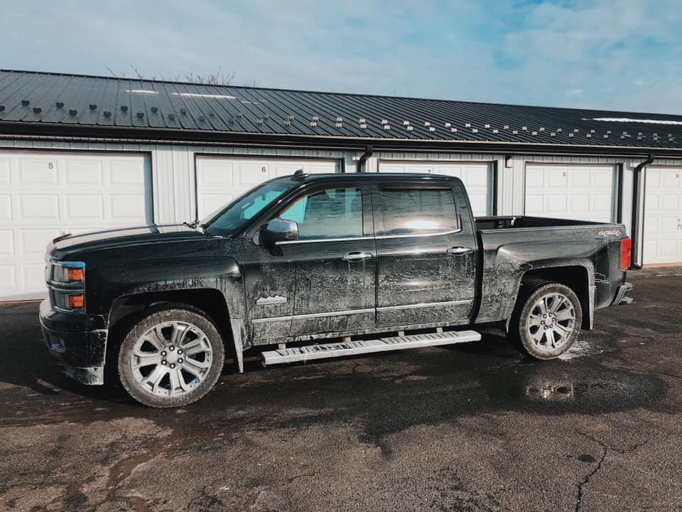 Black pickup truck parked outside garage bays on a wet lot, covered in mud.