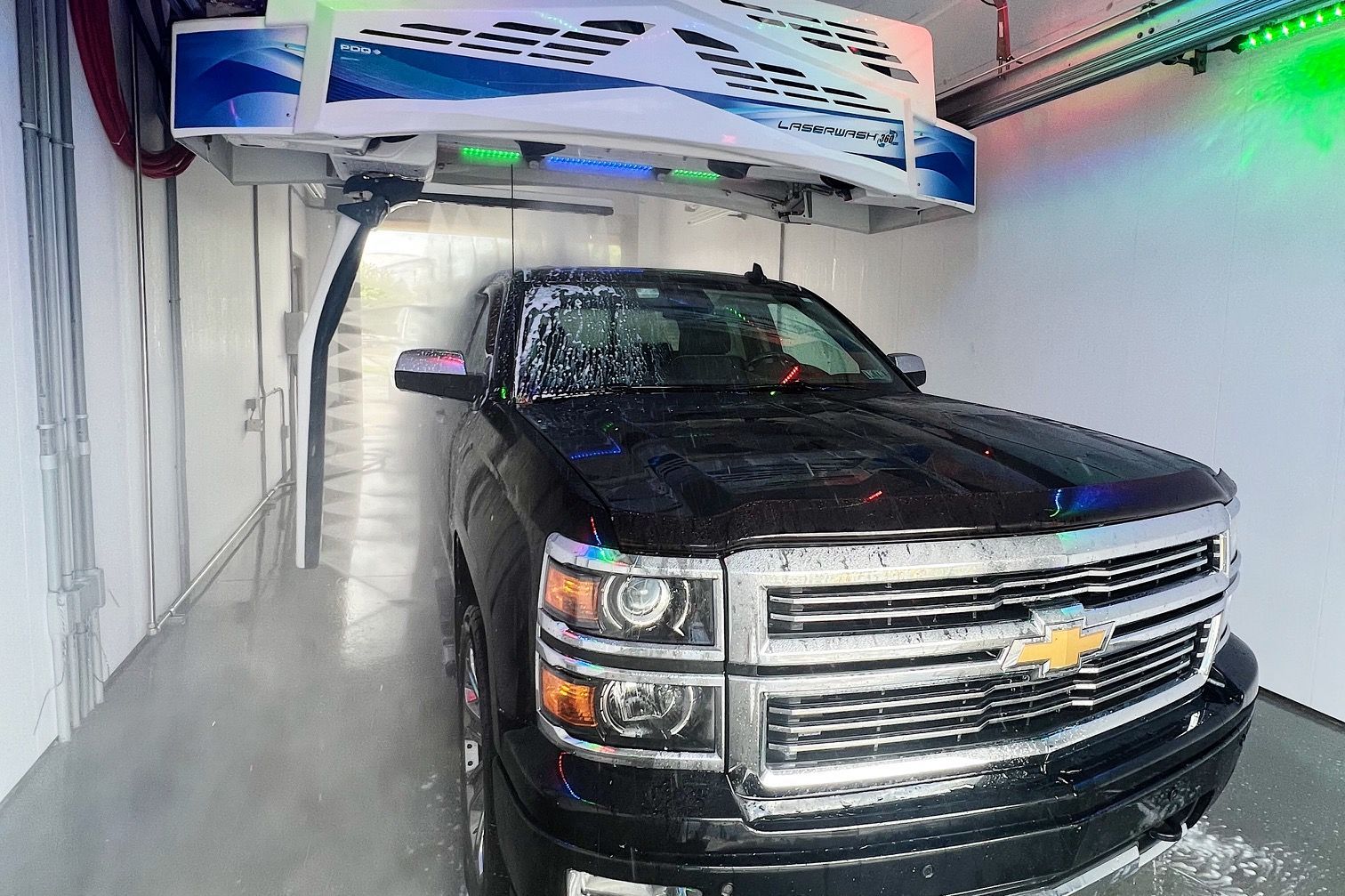 Black Chevrolet pickup in an automatic car wash, surrounded by soap and mist with colorful lights overhead
