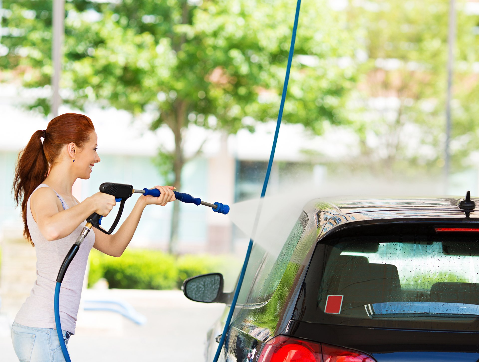 Woman pressure washes the back of a black car outdoors.