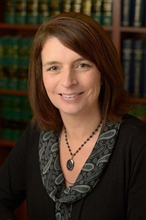 A woman wearing a necklace and a black shirt is smiling in front of a bookshelf. A woman wearing a necklace and a black shirt is smiling in front of a bookshelf.