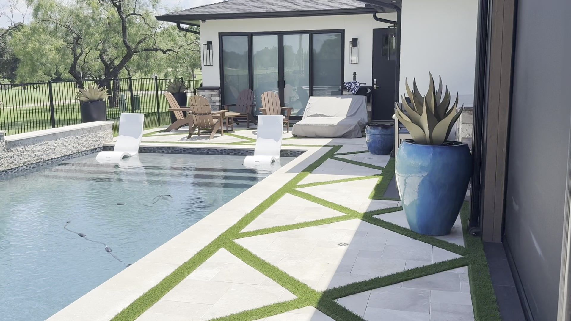 Modern backyard pool with lounge chairs, patio seating, and a potted plant beside a glass-walled house