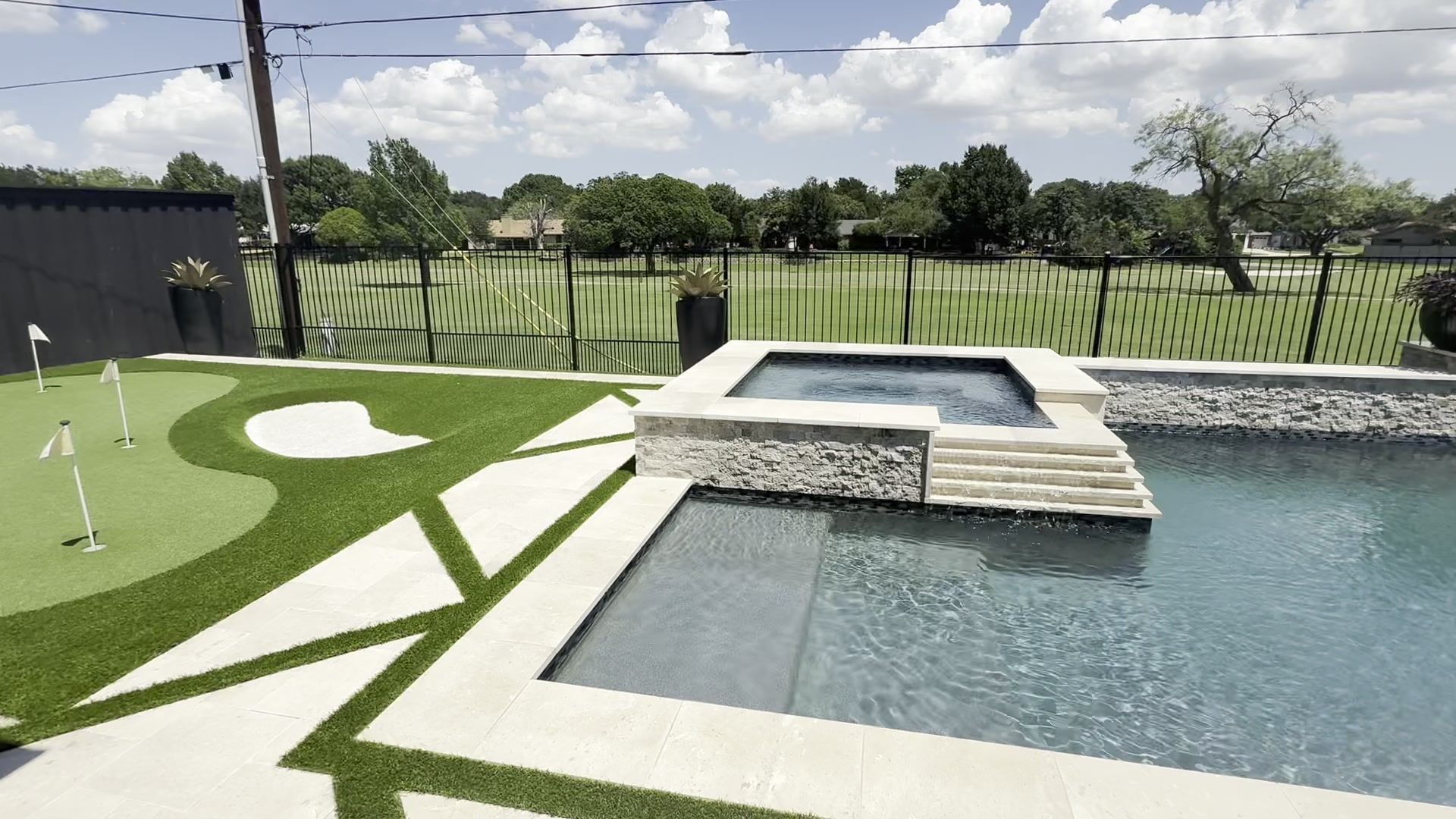 Backyard pool with built-in spa, green turf, and fenced lawn under a partly cloudy sky
