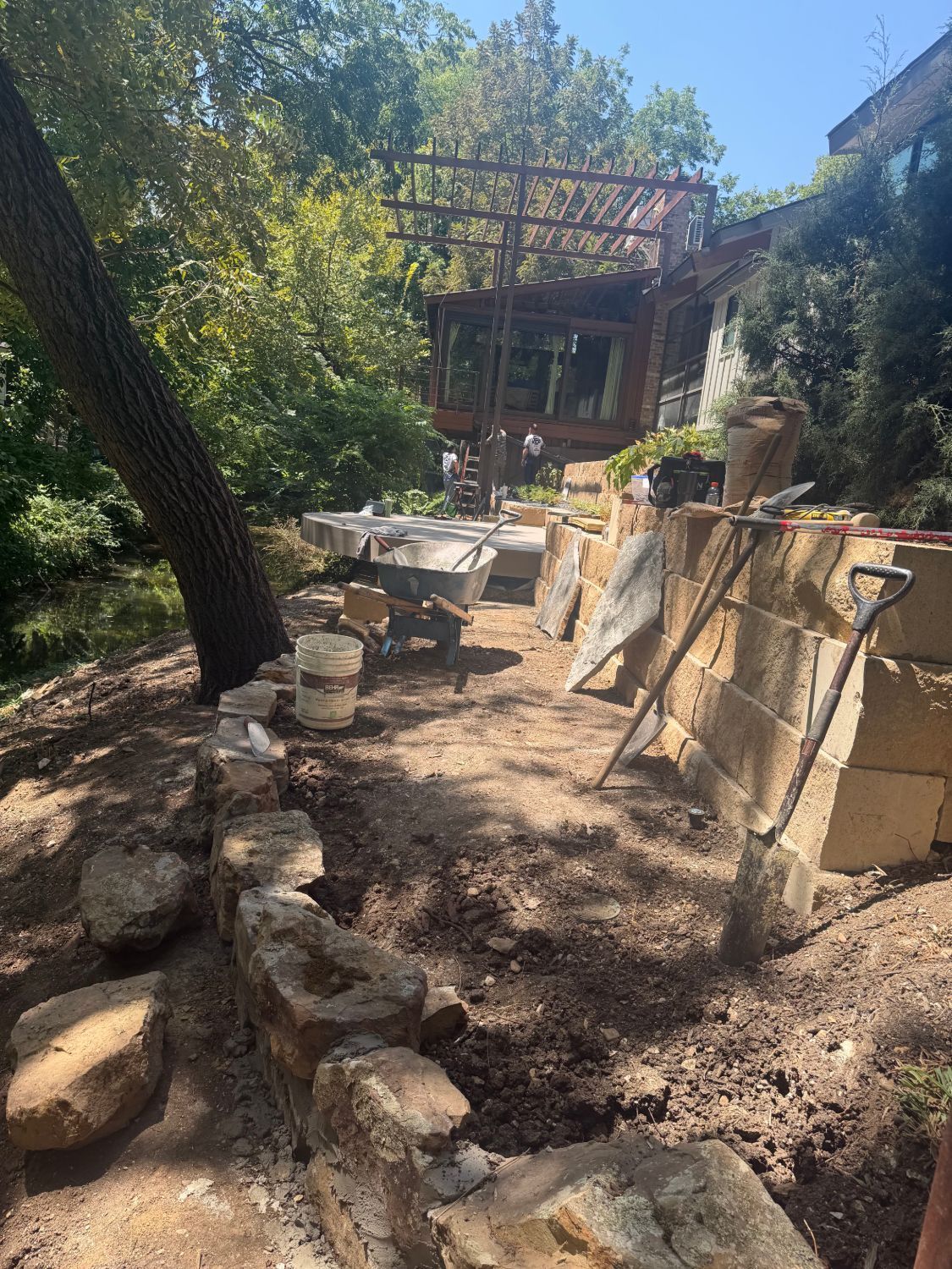 Outdoor stone steps and a dirt path beside a rustic building, with trees and a wooden deck in sunlight