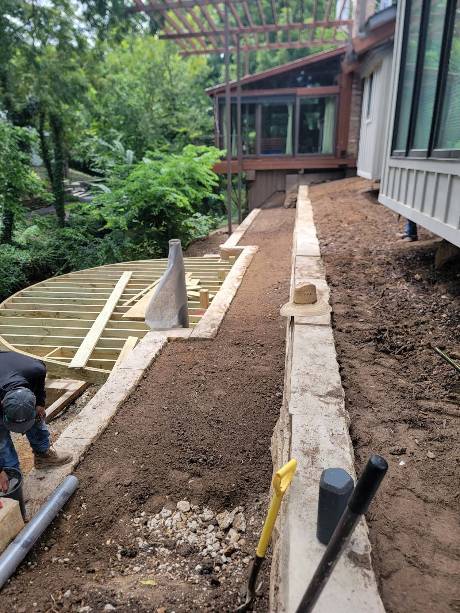 Landscaped walkway under construction beside a house, with concrete edging, gravel, and a worker crouching at left