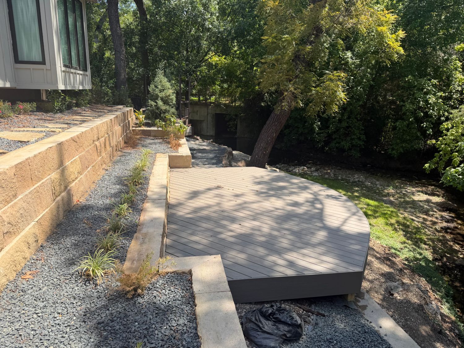 Composite deck and landscaping, bordered by gravel and trees, retaining walls in a shaded yard overlooking a creek in Dallas, Texas.