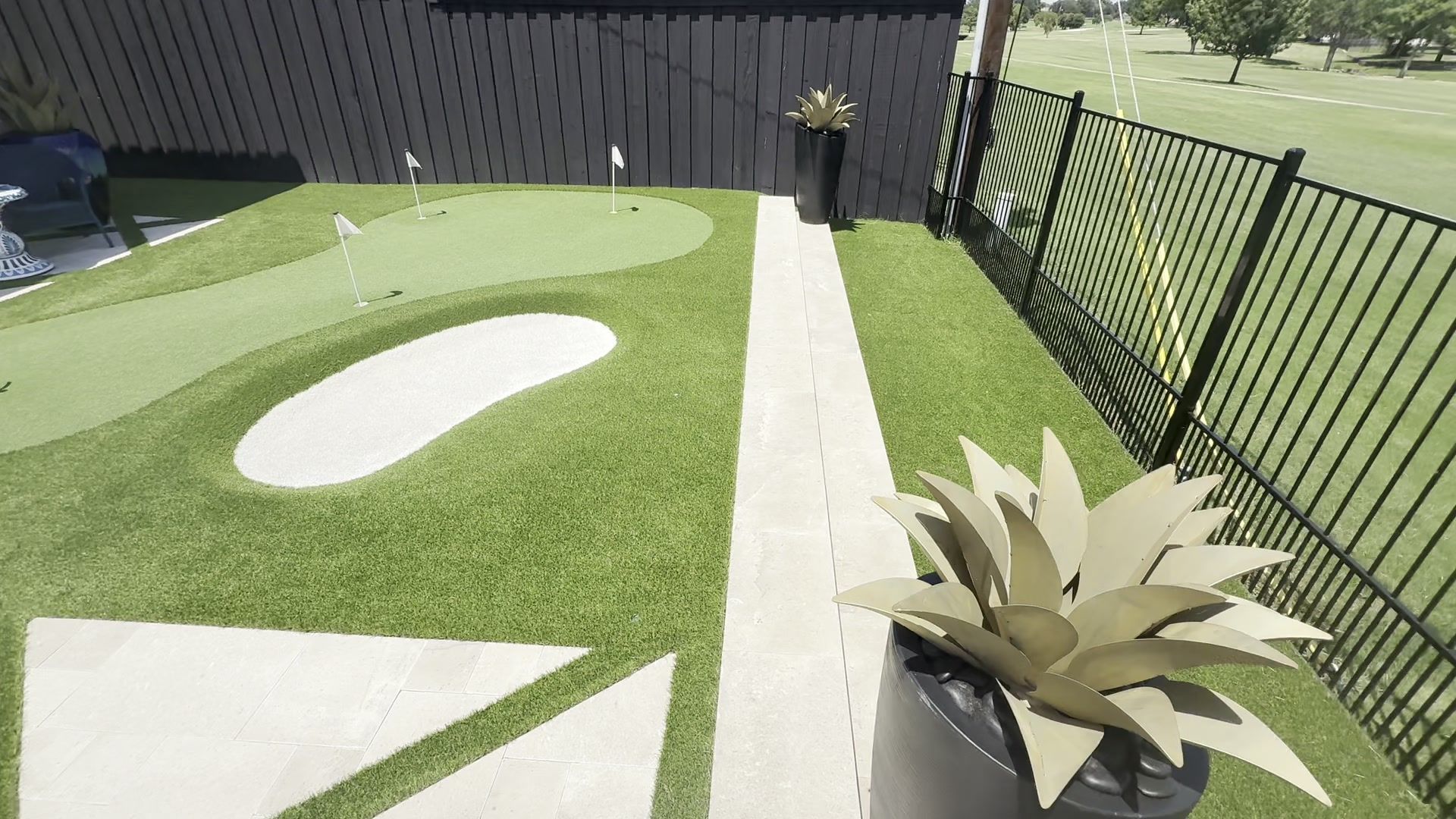 Modern backyard putting green with white sand bunkers, paver path, and potted agave plant alongside a fence