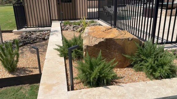 Landscaped rock garden with large boulder, green plants, and concrete edging beside a black metal fence